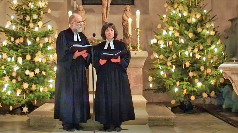 Pfarrerin und Pfarrer Henzler vor Weihnachtsbäumen in der St. Laurentius-Kirche